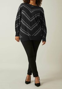 Pull-over noir avec des rayures en forme de chevrons argentés, coupe décontractée, manches longues et poignets côtelés, associé à un pantalon skinny noir et des talons.