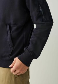 Tumesinine dressipluus soonikkoes varrukate ja alläärega, vasakul varrukal tõmblukuga küljetasku. Sile tekstuur, lõdvestunud istuvus.