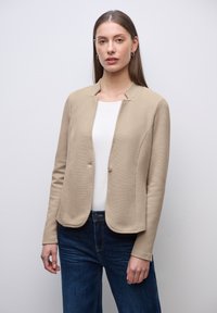 Jeune femme aux cheveux bruns raides, portant une veste beige texturée, une chemise blanche et un jean bleu foncé, debout devant un fond gris clair.