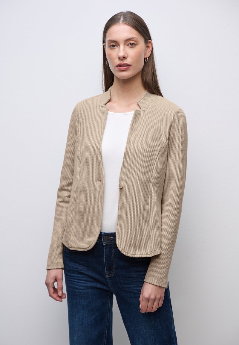 Jeune femme aux cheveux bruns raides, portant une veste beige texturée, une chemise blanche et un jean bleu foncé, debout devant un fond gris clair.
