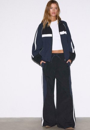 Młoda kobieta z długimi włosami, ubrana w czarno-białą sportową kurtkę, szerokie czarne spodnie dresowe i biały krótki top, stoi na tle białej ściany.