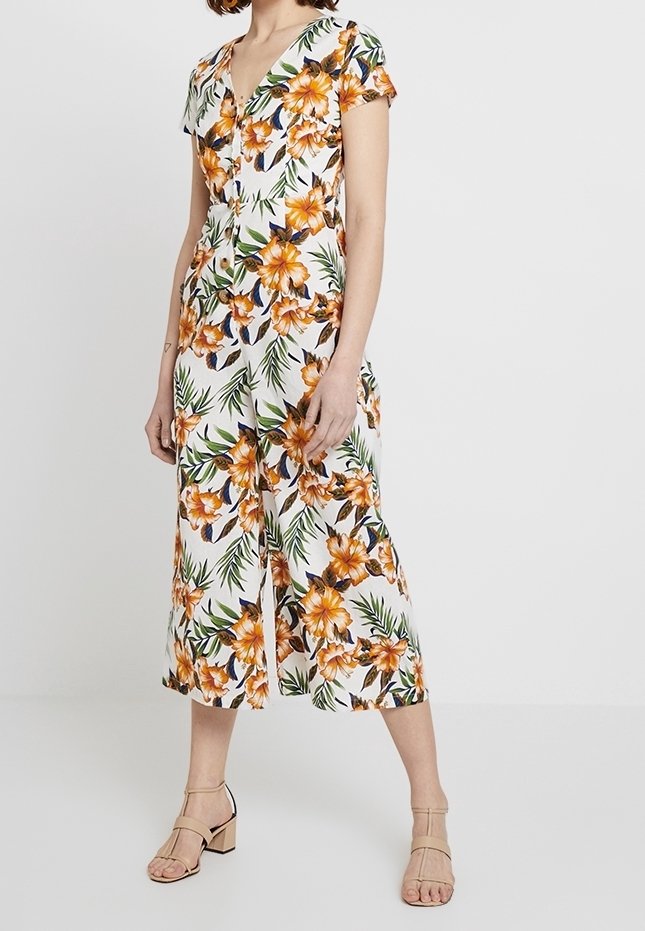 Femme portant une combinaison blanche à manches courtes boutonnée avec imprimé floral orange et feuilles vertes, associée à des sandales beiges à talons blocs.