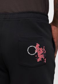 Black sweatpants feature a back pocket adorned with pink floral embroidery and circular text. Made of soft fabric with an elastic waistband.