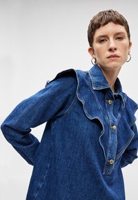 Τζιν πουκάμισο με βολάν στον ώμο, που διαθέτει κλασικό γιακά και δύο μεγάλες χρυσές κουμπότρυπες.