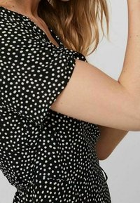 Schwarzes Kleid mit weißen Pünktchen, kurzen fließenden Ärmeln und einer taillierten Taille mit Raffung. Glatte Stoffstruktur.