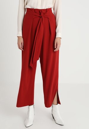 Czerwone szerokie spodnie z wysokim stanem, wiązane z przodu paskiem i z rozcięciami przy kostkach, noszone z białymi botkami na obcasie typu szpic oraz kremową bluzką z długim rękawem.