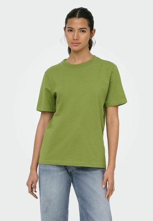 Ung kvinde med mørkt hår og store øreringe iført en ensfarvet grøn kortærmet t-shirt og lyseblå jeans mod en hvid baggrund.