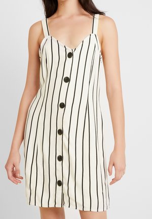 Žena v bílých šatech bez rukávů s vertikálními černými pruhy a šesti černými knoflíky vpředu, na jednobarevném pozadí.