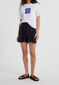 T-shirt en coton blanc avec un texte graphique en carré bleu, associé à une jupe bleu marine foncé et des sandales à brides noires avec une texture tissée.