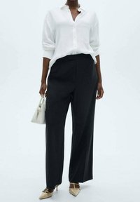 Witte button-up blouse met opgerolde mouwen, gecombineerd met zwarte wijde broek en beige puntige hakken, en houdt een kleine witte handtas vast.