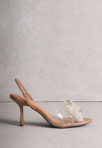 Sandale slingback beige à talon haut avec bride transparente et ornement décoratif en cristal étincelant sur l'orteil.