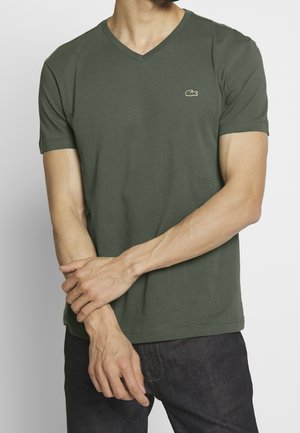 Mann trägt ein dunkelgrünes V-Ausschnitt-T-Shirt mit kurzen Ärmeln und kleinem Krokodil-Logo auf der Brust, dunkle Jeans, steht mit verschränkten Händen.