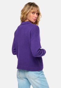 Lila gestrickter Pullover mit Kragen, lockerer Passform, langen Ärmeln und geripptem Saum. Trägt sie mit hellblauen hoch taillierten Jeans.