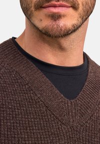 Maglione marrone, texturizzato, con un profondo scollo a V, indossato sopra una camicia nera. Il knit presenta un sottile motivo a coste.