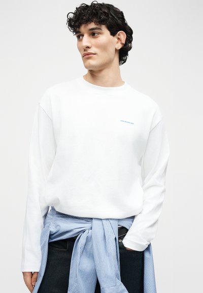 Νέος άνδρας με σγουρά μαύρα μαλλιά, φορώντας λευκό φούτερ με μικρό κείμενο και μπλε ριγέ πουκάμισο δεμένο γύρω από τη μέση του.