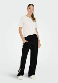 Biały t-shirt z krótkim rękawem w połączeniu z czarnymi spodniami o szerokich nogawkach z elastycznym pasem i bocznymi paskami. Model stoi z ręką w kieszeni.