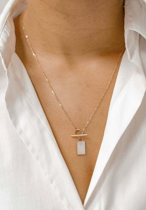 Collana in tonalità oro con un pendente rettangolare dalla superficie liscia e opaca. Stile a catena con chiusura a gancio, indossata su una camicia bianca.