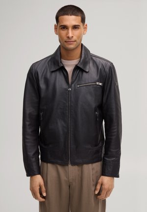 Man die recht vooruit staat, draagt een zwart leren jas met ritssluitingen en beige broek tegen een effen achtergrond.