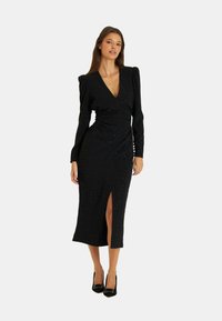 Schwarzes, mittellanges Kleid mit tiefem V-Ausschnitt, langen Puffärmeln und einem seitlichen Schlitz, mit verstreuten Strassdetails. Dazu werden hohe Absätze getragen.