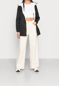 Schwarze wasserdichte Jacke mit weißer Kapuze, Reißverschluss vorne und gelbem Besatz. Kombiniert mit weiten, cremefarbenen Hosen und weißen Turnschuhen.