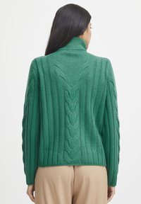 Grüner Kabelstrickpullover mit hohem Kragen, vertikalen Streifen und strukturierten Akzenten entlang des Rückens und der Ärmel.