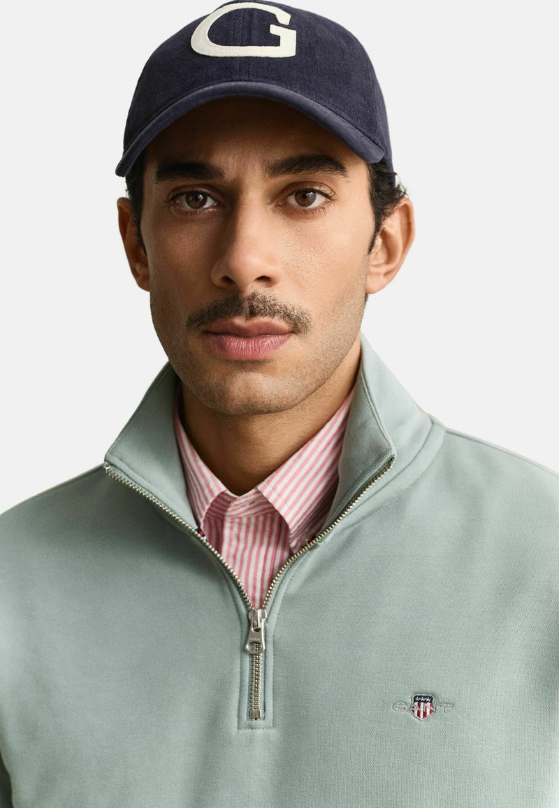 Homme avec moustache portant une casquette marine avec un "G" blanc, un pull à fermeture éclair vert clair et une chemise à rayures roses sur un fond uni.