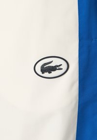 Λευκό ύφασμα με μπλε πλαϊνή ρίγα διαθέτει μια μαύρη οβάλ ετικέτα που απεικονίζει το λογότυπο ενός κροκόδειλου. Το υλικό φαίνεται λείο με μια ελαφριά γυαλάδα.