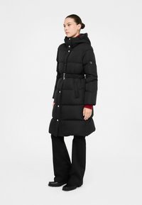 Cappotto piumino nero con vita con cinturino, cappuccio e chiusure a pressione. Tessuto strutturato con un design trapuntato. Abbinato a pantaloni e scarpe nere.