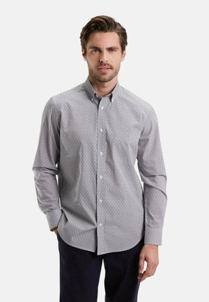 Homme aux cheveux bruns courts portant une chemise à manches longues grise à motifs avec des boutons et un pantalon sombre, se tenant avec une main dans sa poche.