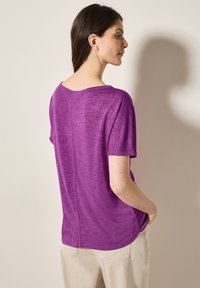 Haut court violet à manches courtes avec un col rond et une coupe décontractée, fabriqué en tissu doux et texturé. Détails de coutures visibles dans le dos.