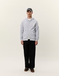 Felpa grigia con zip e tasche frontali, con dettaglio del logo, indossata sopra una maglietta bianca, abbinata a pantaloni neri a gamba larga e a un berretto scuro.