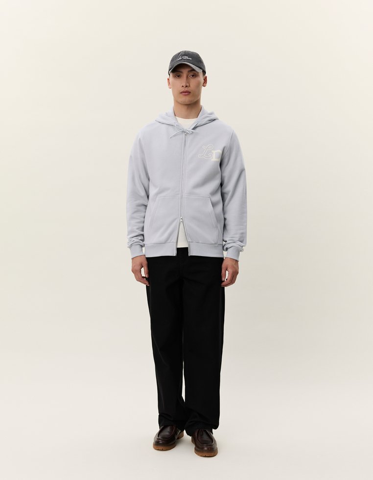 Felpa grigia con zip e tasche frontali, con dettaglio del logo, indossata sopra una maglietta bianca, abbinata a pantaloni neri a gamba larga e a un berretto scuro.