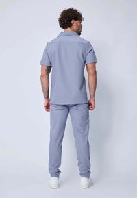 Homme aux cheveux bouclés et tatouages sur les bras, portant une chemise à manches courtes bleu clair et un pantalon assorti, se tenant face à un fond clair uni.