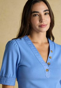 Sieviete ar garajiem tumšajiem matiem, kas valkā zilu ribota V-veida kakla topu ar četriem brūniem pogām un īsām savilkta piedurknēm uz bēša fona.