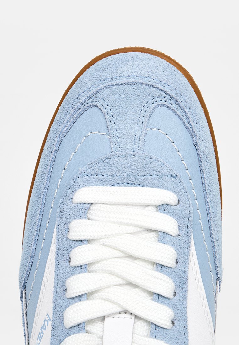 Punta de zapatilla de ante y cuero azul claro con cordones blancos y suela de goma marrón, mostrando costuras y textura detalladas.
