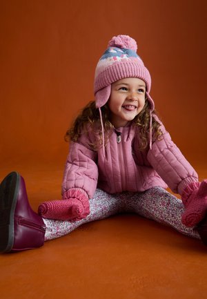 Lachend jong meisje zit op een oranje vloer, draagt een roze winterjas, bloemenlegging, gebreide muts met pompon, wanten en donkerroze laarzen.