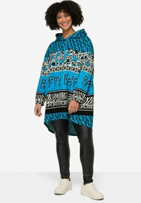 Gebreide poncho in turquoise en zwart, met opvallende patronen en teksten. Lange mouwen, capuchon en een asymmetrische zoom. Gecombineerd met zwarte leggings en witte sneakers.