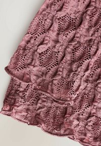 Tissu en dentelle mauve avec des motifs floraux et de feuilles, présentant des formes losangées en ajouré et des bords festonnés, affiché sur un fond clair.