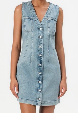 Robe en denim bleu clair sans manches avec fermeture à boutons sur le devant et col en V, portée par une personne montrée du cou jusqu'au milieu de la cuisse.