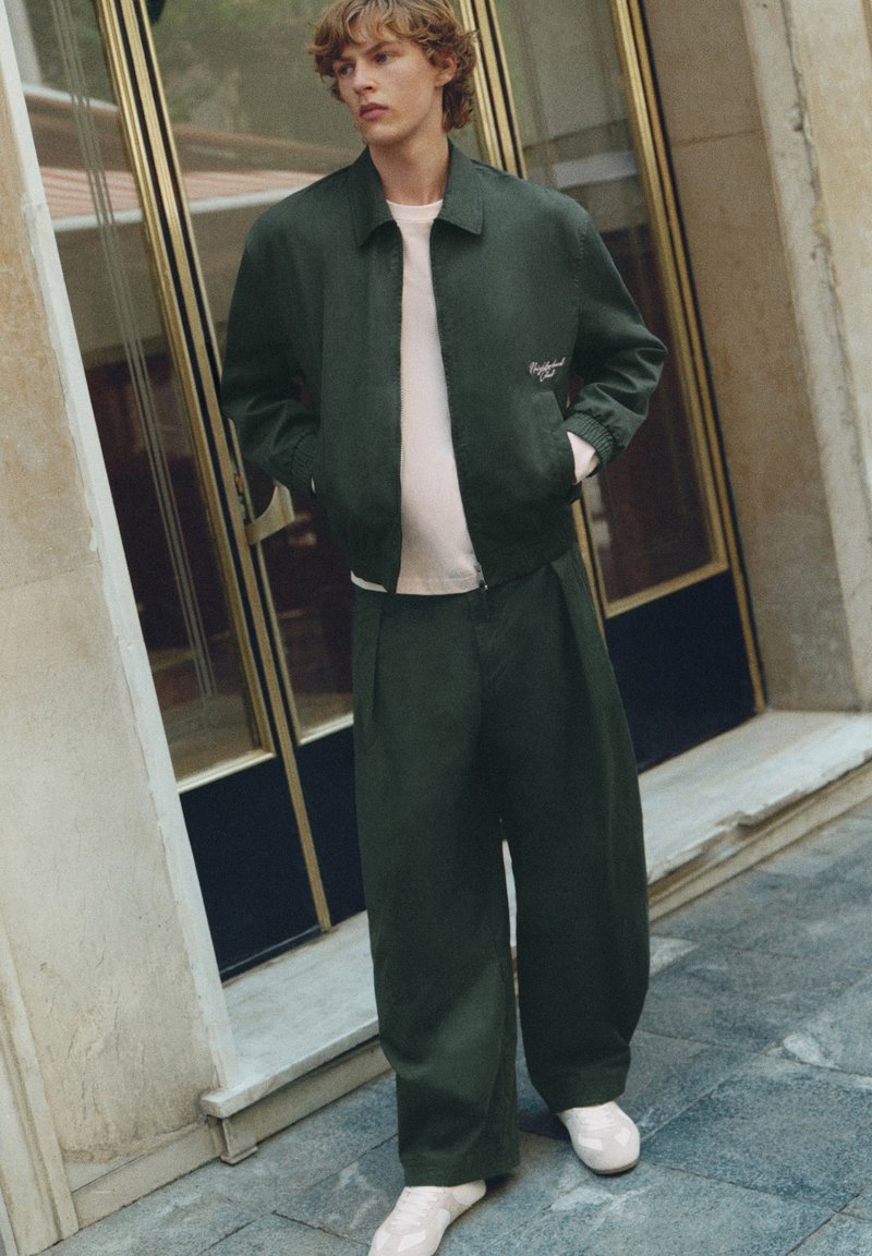 Jeune homme portant un pantalon ample vert foncé et une veste par-dessus une chemise claire, se tenant devant des portes en verre et des murs en pierre.