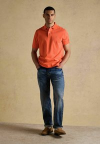 Jeune homme debout les mains dans les poches, portant un polo orange, un jean bleu et des mocassins marron, sur un fond beige uni.