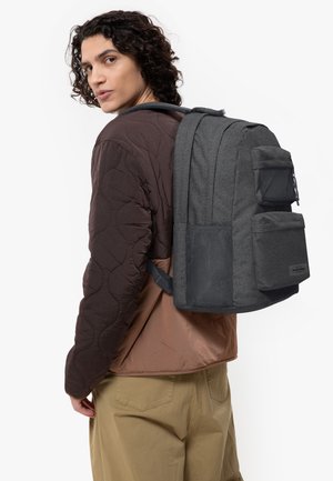 Un sac à dos gris avec plusieurs compartiments et des accents texturés, fabriqué à partir de matériaux durables. Porté sur le dos au-dessus d'une veste marron et d'un pantalon beige.