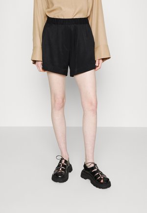 Person iført beige langærmet top, sorte højtaljede shorts og sorte chunky lukkede sandaler står på en grå gulv.