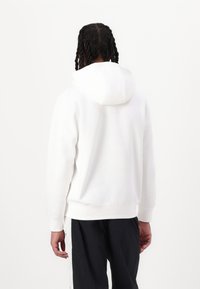 Λευκό φούτερ με κουκούλα και μαλακή υφή, με τσέπη καγκουρό και ριμπ μανσέτες, φαίνεται από πίσω. Φοράει με σκούρο παντελόνι.