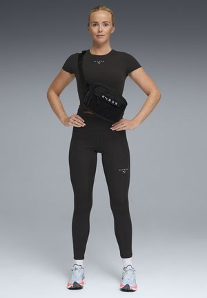 Frau steht mit Händen in Hüfthöhe, trägt schwarze Sportkleidung, weiße Sneakers und eine schwarze Umhängetasche mit der Aufschrift "HYROX" vor grauem Hintergrund.