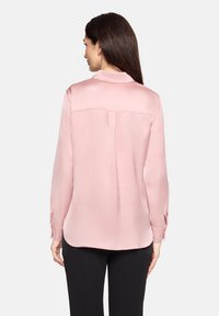 Jasnoróżowa bluzka z długim rękawem, z kołnierzykiem, plisą z tyłu i mankietami na guziki. Wykonana z gładkiej tkaniny, z zaokrąglonym dołem i luźnym krojem.