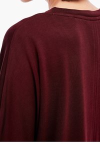 Burgundin neule, jossa on pehmeä, teksturoitu kangas ja löysä istuvuus, tiputetut olkapäät sekä hienovaraiset saumat yksityiskohdat selkäpuolella.