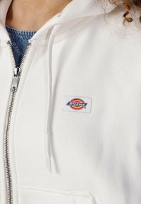 Felpa bianca con zip realizzata in un tessuto morbido, dotata di tasca a marsupio e di una patch ricamata con il logo "Dickies" sul petto.