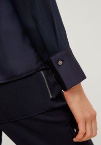 Navy blouse with a smooth fabric, showcasing a buttoned cuff and a contrasting textured panel at the waist with a side zipper detail.
