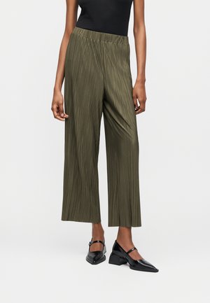 Femme portant un pantalon large plissé vert olive, un haut sans manches noir et des chaussures Mary Jane noires à petit talon, debout sur un fond blanc.